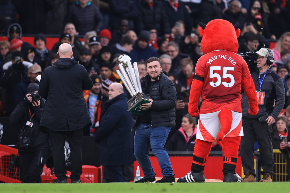Darts-Weltmeister Luke Littler trägt die WM-Trophäe über den Rasen von Old Trafford, daneben das Manchester-United-Maskottchen Fred the Red